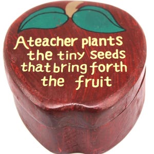 May include: A red wooden apple-shaped box with a green leaf design and the text "A teacher plants the tiny seeds that bring forth the fruit" in yellow. The box is a decorative item.