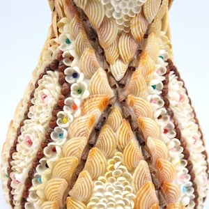 May include: A decorative vase crafted from seashells, featuring a variety of shapes and colors. The vase is adorned with intricate patterns of small, white flower-like embellishments and accents of brown and colorful beads. The overall design is symmetrical and detailed.