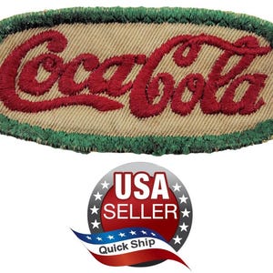 May include: An oval-shaped embroidered patch featuring the Coca-Cola logo in red thread on a tan fabric background, bordered by a thick green stitched edge. The patch has a textured appearance with visible thread work throughout the design.
