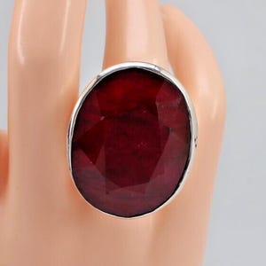 Op de afbeelding: Een grote ovale ring met een dieprode, gefacetteerde edelsteen gezet in een gepolijste zilverkleurige metalen band. De steen vertoont een donkere karmozijnrode tint met zichtbare interne facetten en een glad, reflecterend oppervlak.