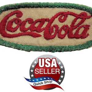 May include: An oval-shaped embroidered patch featuring the Coca-Cola logo in red thread on a tan fabric background. The patch is bordered by a thick, textured green stitched edge. The design is centered and clearly legible against the neutral base.