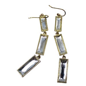 May include: A pair of drop earrings featuring three rectangular clear crystals set in gold-toned metal frames. Each earring consists of three vertically linked segments that hang from a curved ear wire, creating a linear and articulated design.