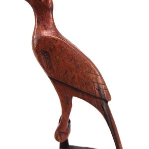May include: A hand-carved wooden bird figurine featuring a dark brown finish with visible wood grain. The bird has a long, curved beak, a crest on its head, and a single bright red eye. It stands on a triangular base with a support strut.