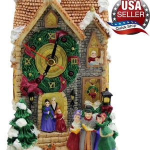 May include: A decorative Christmas clock shaped like a snow-covered house with a wreath-adorned clock face. The clock features a detailed facade with figurines of carolers and families, and a small Christmas tree. The clock measures approximately 8 inches tall.