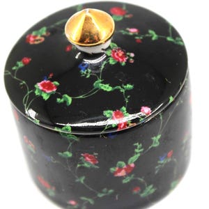 May include: A cylindrical black ceramic jar with a matching lid, featuring a repeating pattern of small red flowers and green vines. The lid is topped with a smooth, conical gold-colored knob, providing a contrast to the dark floral surface.