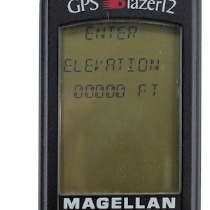 Könnte beinhalten: Ein schwarzes, tragbares Magellan GPS Blazer12-Gerät mit einem digitalen Bildschirm, der den Text ENTER, ELEVATION und 00000 FT anzeigt. Das Gerät verfügt über ein Steuerkreuz, Navigationstasten wie NAV, GOTO, MARK, MENU, PWR und ein rotes USA SELLER-Abzeichen.