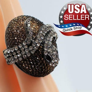 May include: A statement ring featuring a large, oval-shaped centerpiece covered in dark brown crystals. A coiled snake figure, encrusted with clear white crystals and featuring black eyes, wraps around the top of the brown crystal base.