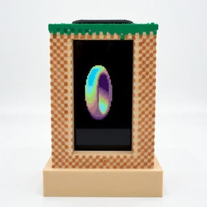 Pode incluir: Uma moldura de ecrã digital retangular com um padrão de xadrez bege e branco pixelizado e uma borda superior verde. O ecrã mostra um anel de pixel art multicolorido sobre um fundo preto, assente numa base bege sólida.