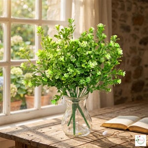Puede incluir: Un ramo de plantas artificiales verdes con pequeñas flores blancas en un jarrón de cristal transparente. El jarrón está sobre una mesa de madera cerca de un libro abierto y gafas, con una ventana al fondo.
