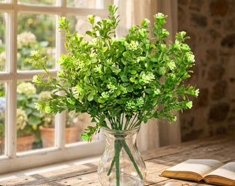May include: A bunch of artificial green plants with tiny white flowers in a clear glass vase. The vase is on a wooden table near an open book and spectacles, with a window in the background.