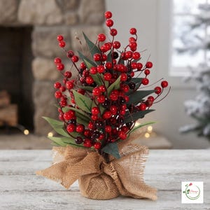 May include: A decorative arrangement featuring numerous small, glossy red berries on thin brown stems interspersed with pointed green leaves. The base of the arrangement is wrapped in a textured, light brown hessian fabric tied with a bow.
