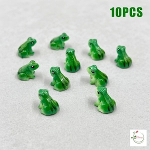 Könnte beinhalten: Ein Set aus 10 kleinen grünen Froschfiguren, jede mit einer glatten, glänzenden Oberfläche, dunkelgrünen Streifen auf dem Rücken und schwarzen Augen. Der Text 10PCS ist in der oberen rechten Ecke des Bildes zu sehen.