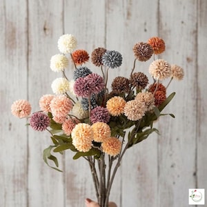 Pode incluir: Um buquê de flores artificiais com flores esféricas em várias cores, incluindo pêssego, laranja, creme e roxo. Os caules são marrons e as folhas são verdes. O fundo é uma superfície de madeira clara.