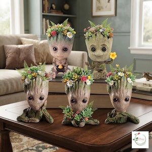 Peut inclure: Un ensemble de cinq jardinières en forme de personnage arboricole avec des surfaces texturées rappelant l'écorce, chacune dotée de grands yeux sombres et remplie de succulentes artificielles, de petites fougères et de fleurs colorées.