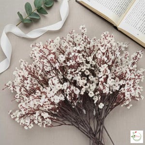 Può includere: Un mazzo di fiori artificiali bianchi con steli e rami marrone scuro. I fiori sono raggruppati, creando un aspetto pieno e strutturato. Sono presenti anche un nastro bianco e un rametto di foglie verdi.