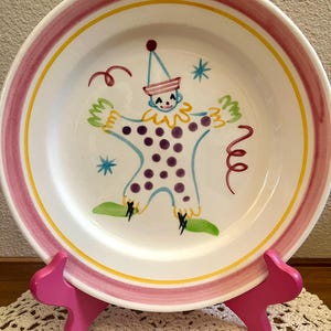 May include: A white ceramic plate featuring a hand-painted clown illustration in the center. The clown has purple polka dots, green shoes, and a blue hat. The plate has a wide pink rim with a thin yellow border. It rests on a bright pink stand.