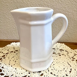 May include: A white ceramic pitcher with a smooth, glossy finish and a distinct hexagonal body shape. The pitcher features a curved handle on the right side and a wide, pointed pouring spout at the top, showcasing a clean and simple design.