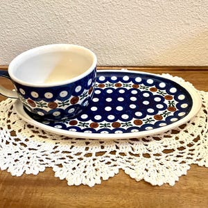 May include: A ceramic cup and matching oval saucer featuring a dark blue background with a repeating pattern of white circles, brown dots, and small green leaf motifs. The interior of the cup is white, and the set has a glossy glazed finish.