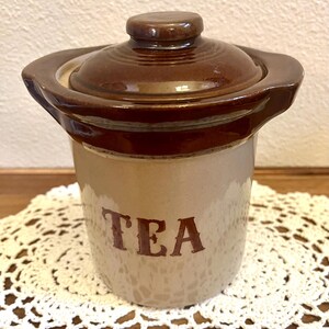 May include: A cylindrical ceramic tea canister featuring a two-tone design with a light beige base and a dark brown glazed rim and lid. The word TEA is printed in dark brown capital letters on the front. It has two small side handles.