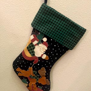 May include: A Christmas stocking featuring a black fabric body with white polka dots and a green and black buffalo check cuff. The stocking is decorated with fabric appliques of Santa Claus, a reindeer, and gift boxes.