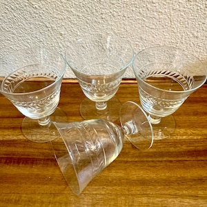 May include: Four clear glass goblets with short stems and circular bases. Three of the glasses are standing upright, while one is lying on its side. Each glass features a decorative etched band of repeating leaf-like patterns around the upper bowl.