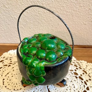 May include: A black cauldron with a metallic handle and three small feet. The top is filled with sculpted, glossy green bubbles that spill over the front rim. The surface of the cauldron has a dark, matte finish with subtle texture.