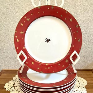 May include: A stack of round ceramic plates featuring a wide red rim decorated with gold-colored star patterns of varying sizes. The center of each plate is white, with a single small, dark, star-shaped icon printed in the very middle of the surface.