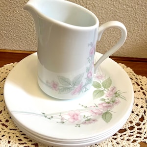 May include: A white ceramic creamer pitcher and a stack of matching white plates, each decorated with a delicate pattern of soft pink flowers and muted green leaves, featuring a curved handle and a pouring spout on the creamer.