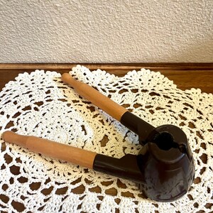May include: A nutcracker featuring two light brown wooden handles attached to a dark brown, rounded metal crushing mechanism. The tool has a hinged design with a central circular opening for placing nuts, and the handles extend outward.