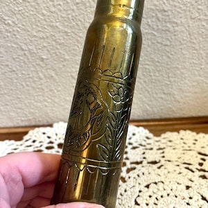 May include: A cylindrical brass casing featuring an engraved floral pattern with scalloped borders. The metal surface has a polished, reflective gold-toned finish with visible etched details of flowers and leaves wrapping around the central body.