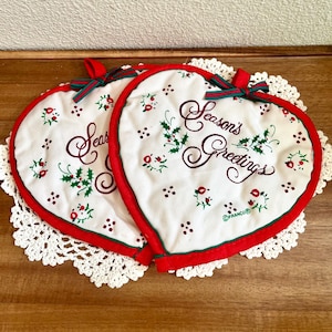 May include: Two heart-shaped white fabric pot holders with red trim and a green and red ribbon bow at the top. The fabric features a pattern of small red berries and green holly leaves, with the text Season's Greetings printed in dark red script.