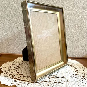 May include: A rectangular picture frame with a metallic gold finish and a decorative embossed pattern along the outer edge. The frame features a smooth, beveled inner border and a clear glass front protecting a light beige, textured paper insert.