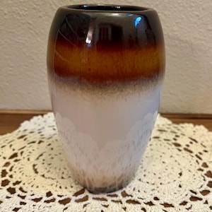 May include: A ceramic vase with a smooth, rounded silhouette featuring a gradient color scheme. The top rim is a deep, glossy dark brown that transitions into a warm amber band, fading into a creamy white base with a subtle, mottled texture.