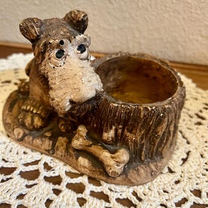 May include: A brown and beige dog figurine featuring a textured, tree-stump-shaped hollow container on its side. The dog has dark eyes, a prominent snout, and is positioned next to a small bone-shaped object on a flat, oval-shaped base.