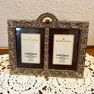 May include: A gold-plated double picture frame by Philip Whitney Ltd. featuring an ornate, embossed leaf pattern and a central oval accent. The frame holds two 2 inch by 3 inch photos and includes text stating Gold-Plated Frame, Hand polished and lacquered, tarnish-resistant.