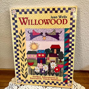 May include: A rectangular book titled Willowood by Jean Wells, featuring a colorful quilt-style cover illustration of houses, flowers, a sun, and two flying figures. The border is a checkered pattern with small flowers and triangular shapes.