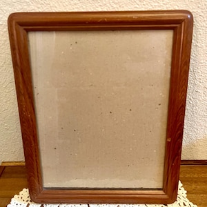 May include: A rectangular picture frame with a medium-brown wood grain finish and rounded corners. The frame features a smooth, polished surface and contains a plain, light-colored backing board visible through the center opening.