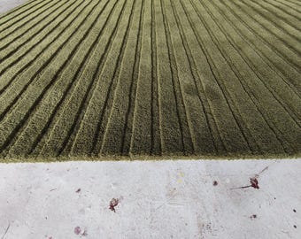 Puede incluir: Una alfombra rectangular de color verde oliva intenso con una superficie texturizada compuesta por rayas verticales paralelas en relieve que recorren toda la longitud de la pieza, creando un patrón lineal constante en todo el diseño.