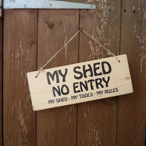 Peut inclure: Panneau rectangulaire en bois clair avec l'inscription noire "MY SHED NO ENTRY" suspendu à une porte en bois marron. Le texte "MY SHED - MY TOOLS - MY RULES" est également visible. Le panneau est suspendu par une ficelle.