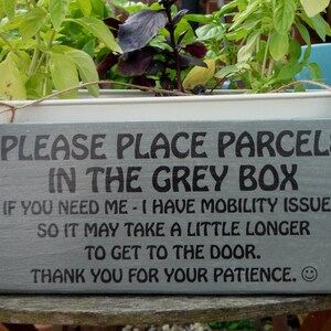 May include: A gray rectangular sign with black text that reads "PLEASE PLACE PARCELS IN THE GREY BOX." The sign also includes a message about mobility issues and a thank you. The sign is hanging in front of a planter box with green plants.