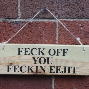 May include: A rectangular light-coloured wooden sign with a natural grain texture, featuring the black text "FECK OFF YOU FECKIN EEJIT" printed in a bold, sans-serif font. The sign is suspended by a thin, light-coloured twine string attached to the top.