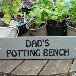 May include: A gray wooden sign with the text "DAD'S POTTING BENCH" in black lettering. The sign is placed in front of various potted plants, including basil and other herbs, on a wooden table.