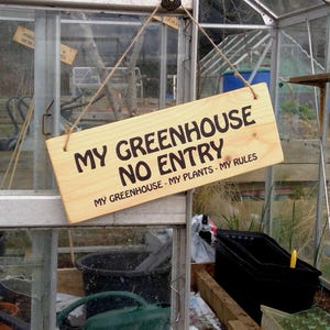 May include: A wooden sign with the text "MY GREENHOUSE NO ENTRY" hangs in a greenhouse. The sign also reads "MY GREENHOUSE - MY PLANTS - MY RULES". The greenhouse has glass panels and contains plants and gardening supplies.