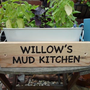 May include: A rectangular light-coloured wooden sign with the text WILLOW'S MUD KITCHEN burnt into the surface in a dark font. The sign features two small pre-drilled holes on the left and right sides for mounting purposes and has a smooth finish.