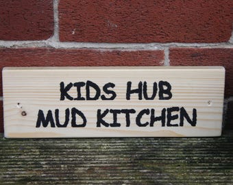 May include: A rectangular light-coloured wooden sign featuring the black text "KIDS HUB MUD KITCHEN" printed in a bold, sans-serif font. The sign has two small pre-drilled holes on the left and right sides for mounting purposes.