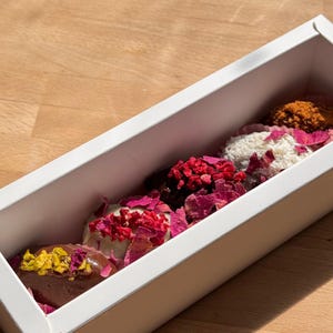 May include: A white rectangular box containing a selection of gourmet chocolates. The chocolates are adorned with toppings such as dried rose petals, coconut flakes, and crushed berries. The box rests on a wooden surface.