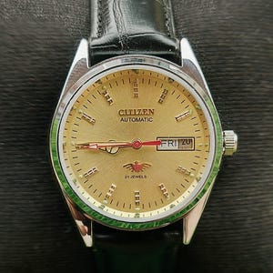 Pode incluir: Um relógio de pulso automático Citizen dourado com uma pulseira de couro preta. O mostrador mostra o dia e a data, com o texto "Citizen Automatic" e "21 Jewels". O relógio tem uma moldura prateada e um anel interno verde.