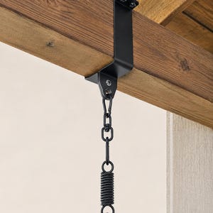 May include: Black metal swing hanger attached to a wooden beam. The hanger includes a chain, spring, and carabiner clip for secure suspension. The wooden beam has a natural wood grain.