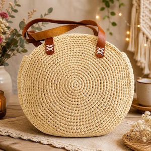 May include: A round, beige crochet handbag with brown leather handles. The bag features a spiral pattern and is accented with brown leather details at the handle attachments. The bag is a light beige color and has a natural, handmade aesthetic.