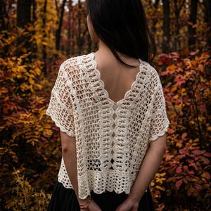 May include: An off-white crochet top featuring a V-neckline, short sleeves, and an intricate openwork pattern with scalloped edges. The garment has a loose, boxy fit and a textured, hand-crafted appearance with a repeating geometric design.